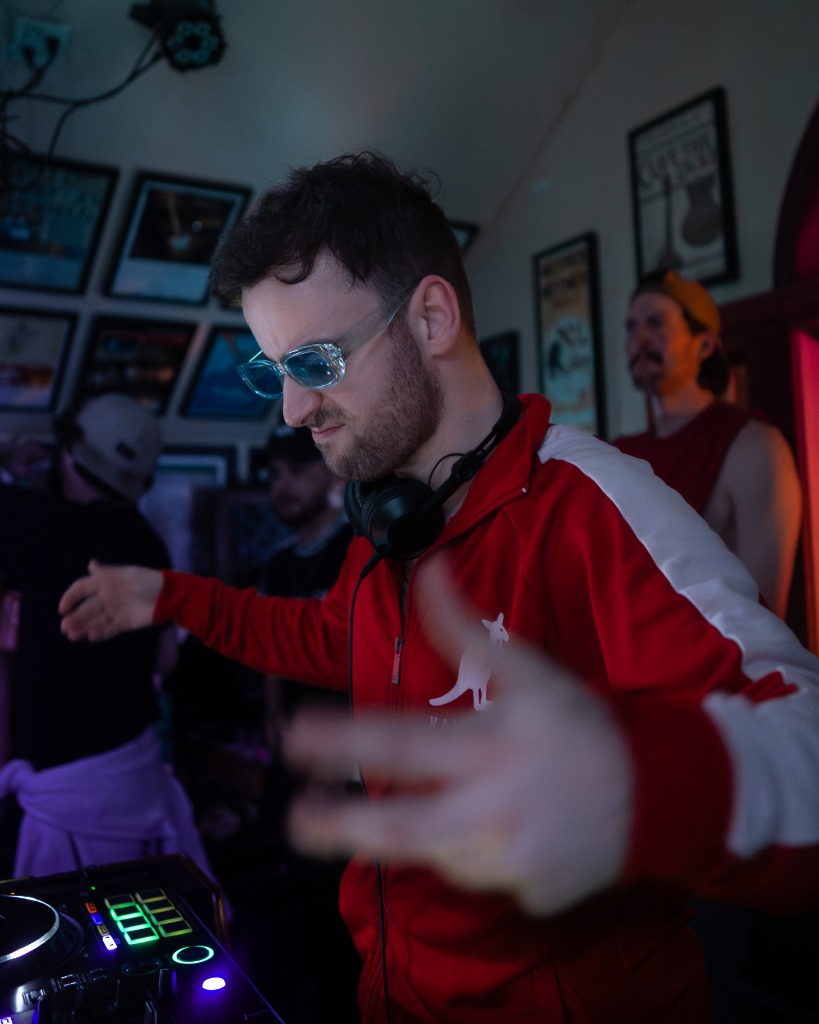 DJ in red track jacket and sunglasses gesturing under blue spotlights at a PEI music gig - Eastside Photography artist promo